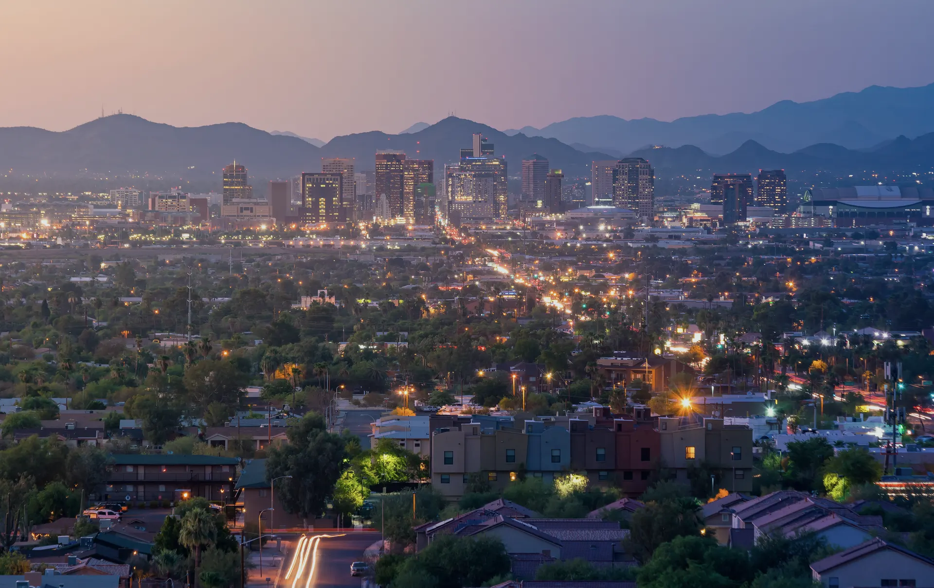 Downtown Phoenix at dusk, city lights on, mountains behind.